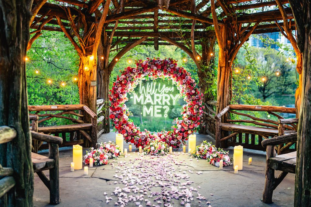 Ring of red and pink flowers with a neon Will You Marry Me sign in a Central Park gazebo, petals along the aisle