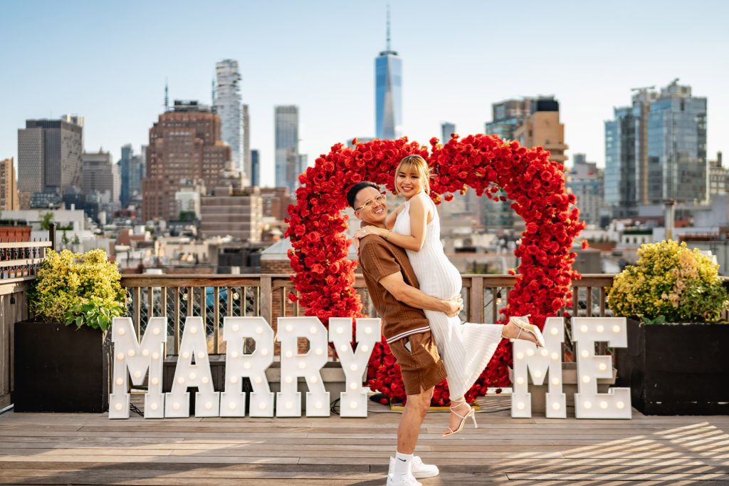 Rooftop proposal downtown with a red rose heart and MARRY ME letters, One World Trade Center behind