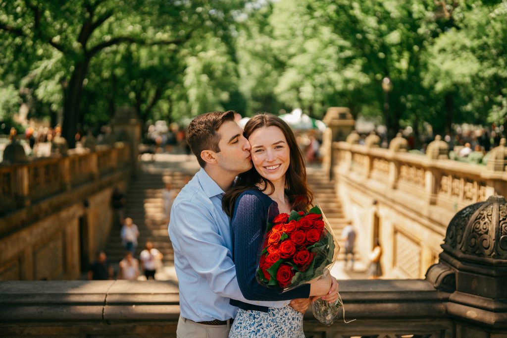 Newly engaged couple kissing with a bouquet of red roses on a Central Park bridge, visitors in the background