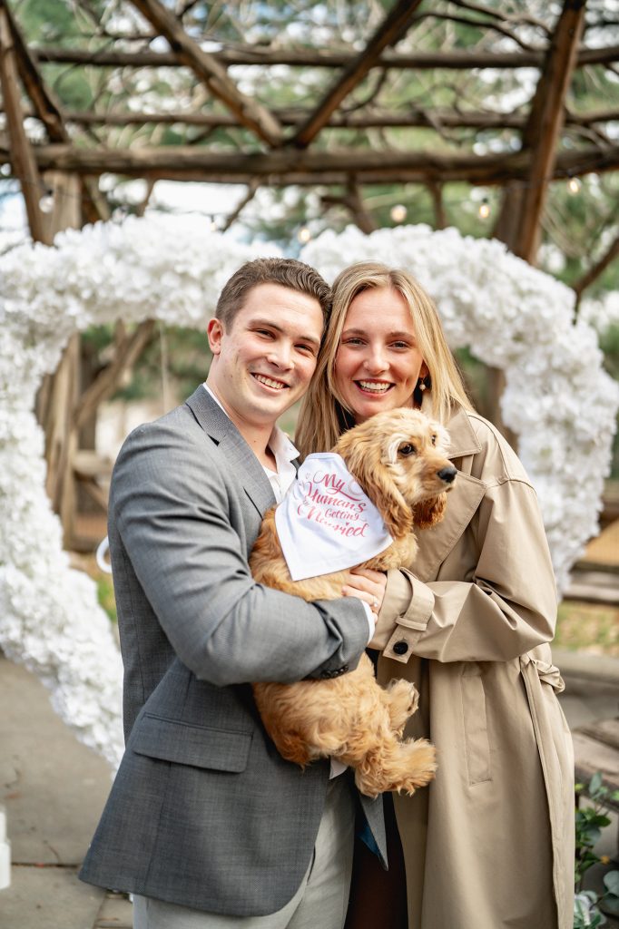 Newly engaged couple holding their dog under a white floral arch at Cop Cot, Central Park