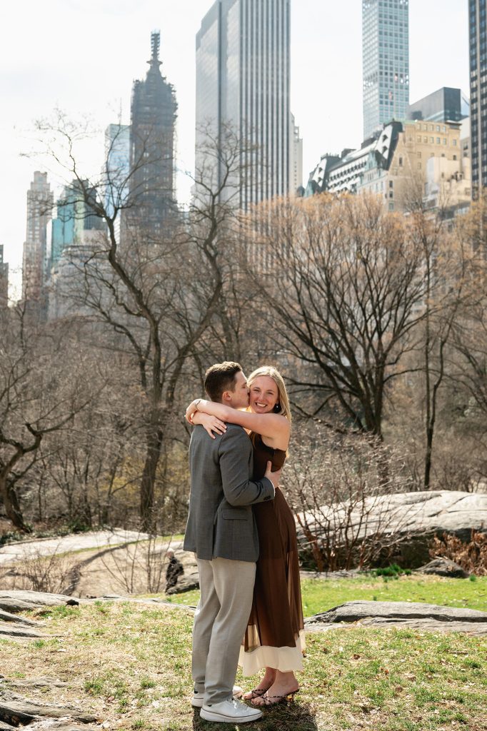 Newly engaged couple embracing in Central Park with the Midtown skyline behind the bare trees