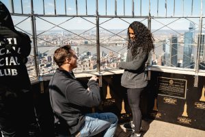 Proposal on the Empire State Building observation deck: man on one knee at the steel cage rail, the city far below, clear blue sky