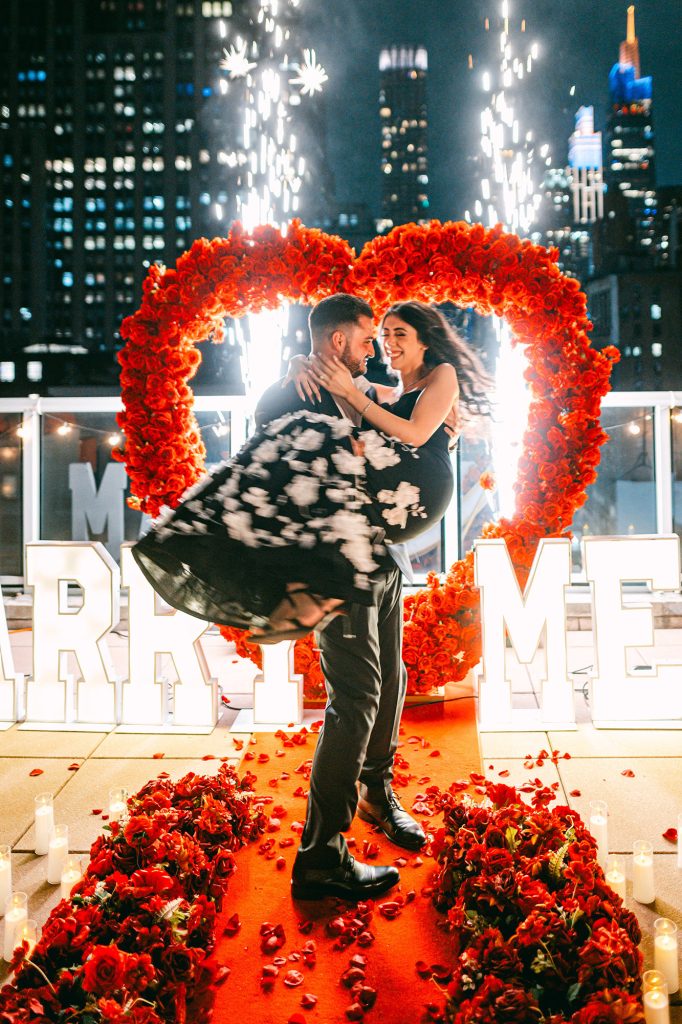 Night rooftop proposal in New York: couple lifted inside a red rose heart arch as sparkler fountains fire