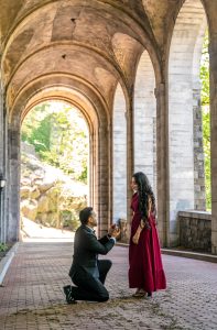 Proposal at Fort Tryon Park: man on one knee inside a Romanesque stone arch, woman in a red gown, greenery beyond the opening