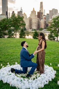 Proposal at Four Freedoms Park on Roosevelt Island: man on one knee inside a ring of white flowers, candles around it, the Midtown skyline across the East River behind them