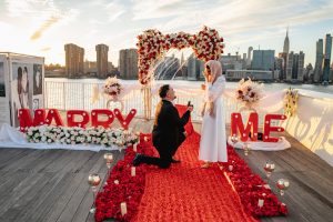 Evening proposal in Long Island City: red rose carpet, floral arch with a fountain and giant MARRY ME letters, man on one knee, woman in white