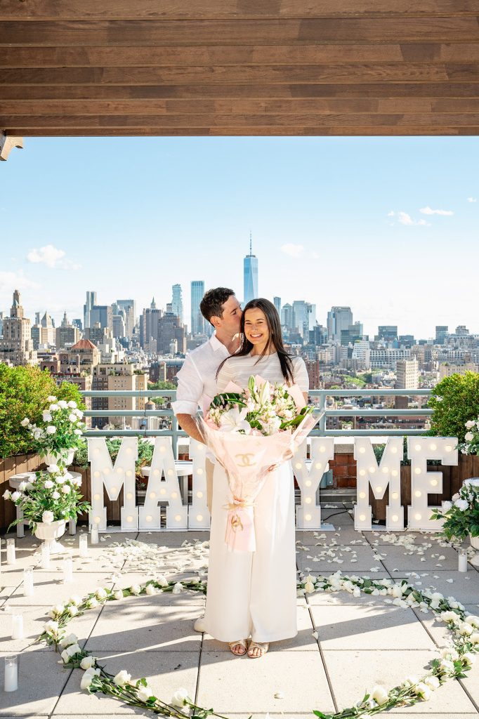 Woman holding a white bouquet beside MARRY ME letters on a private Manhattan rooftop
