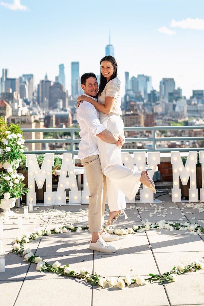 Man lifting his fiancee inside a white flower heart on a Manhattan rooftop, One World Trade Center behind