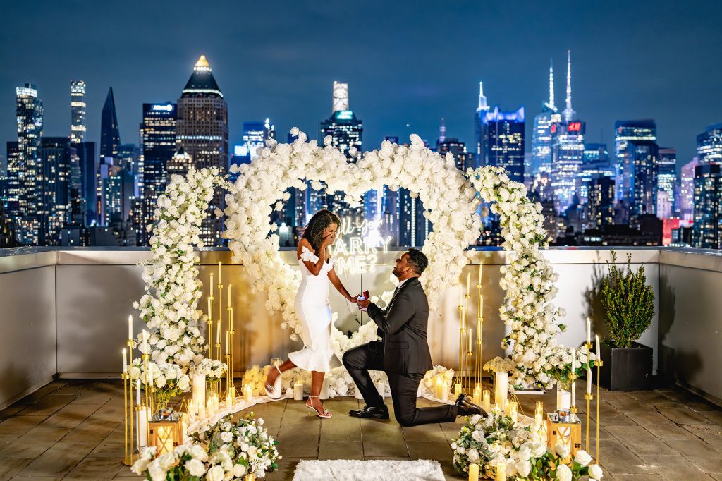 Night rooftop proposal at Midtown View Rooftop Terrace in Manhattan under a white floral arch, city skyline behind