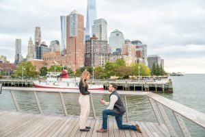 Proposal on Pier 26 with Lower Manhattan and a historic fireboat behind, man on one knee on the boardwalk