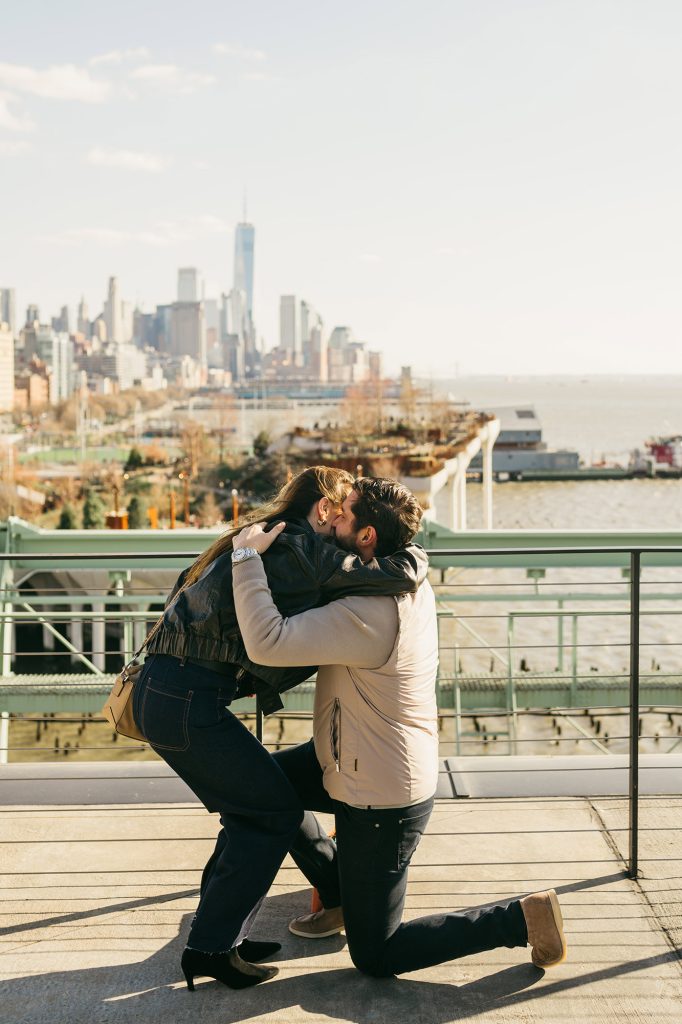 Woman hugging her partner as he kneels at Pier 57 in Manhattan, the Hudson River behind