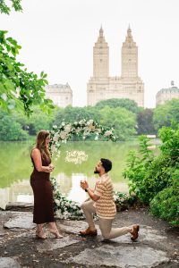 Surprise proposal under a white floral arch at San Remo View in Central Park, San Remo towers across the Lake