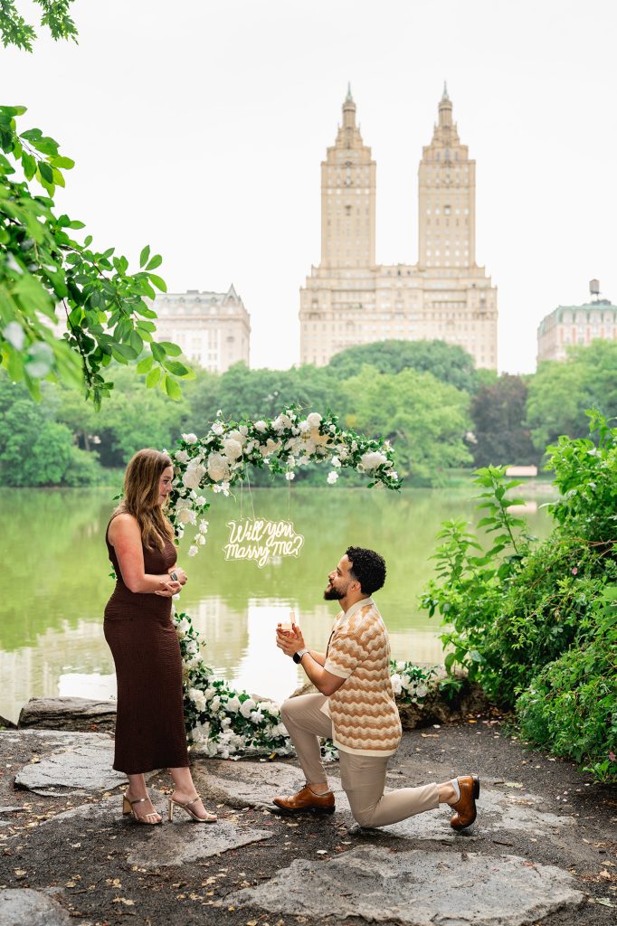 Surprise proposal under a white floral arch at San Remo View in Central Park, San Remo towers across the Lake