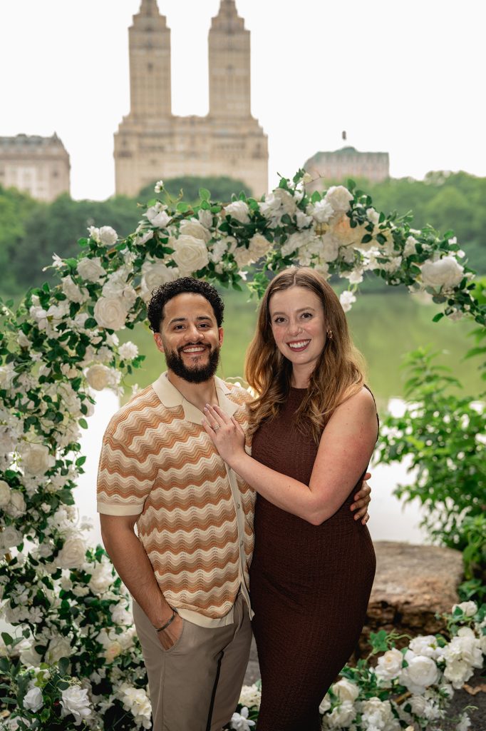 Newly engaged couple under a white floral arch by the Lake in Central Park, San Remo towers behind
