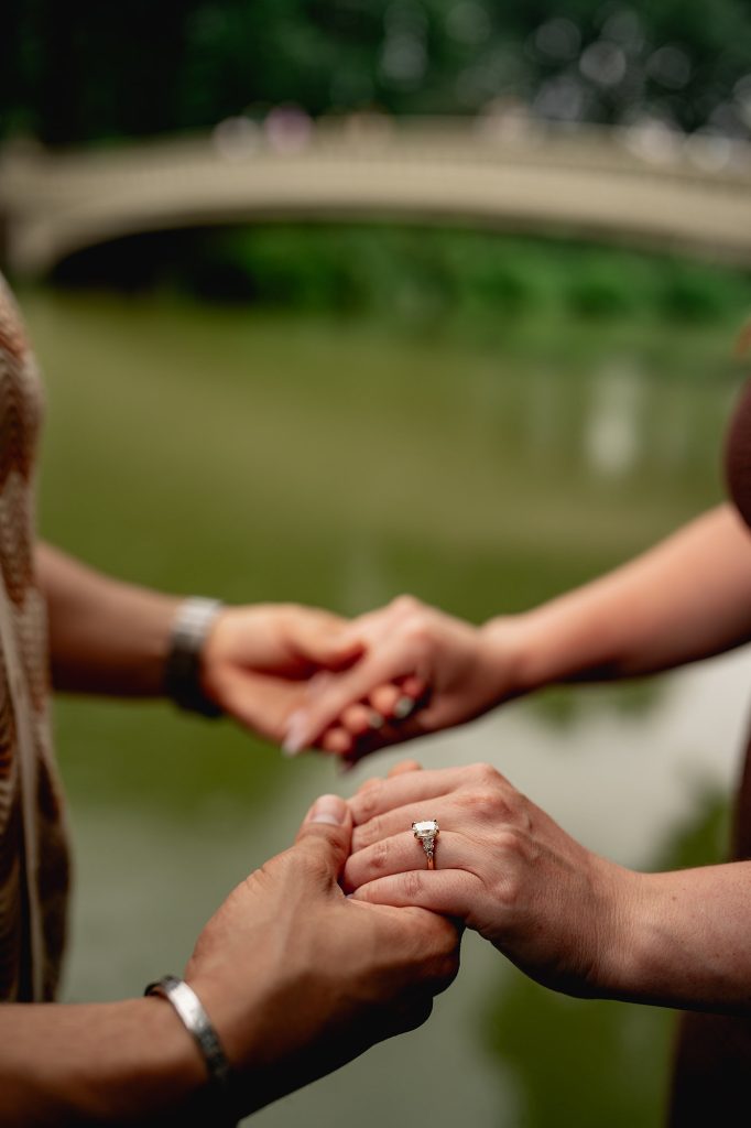 Close-up of an engagement ring in Central Park with Bow Bridge out of focus behind