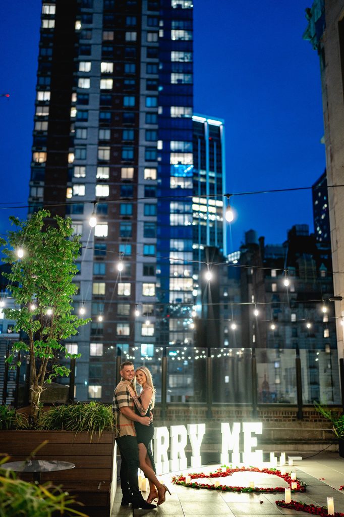 Newly engaged couple beside lit MARRY ME letters on a Midtown rooftop in New York at night