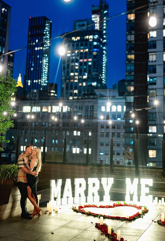 Lit MARRY ME letters and a red rose heart on a Midtown Manhattan rooftop at blue hour