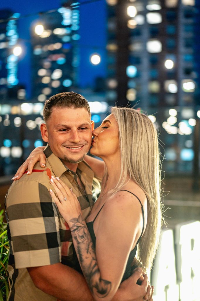 Woman kissing her fiance on a Midtown Manhattan rooftop with city lights behind them