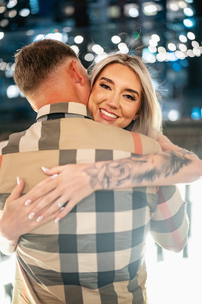 Newly engaged couple on a Midtown rooftop at night, her engagement ring on his shoulder