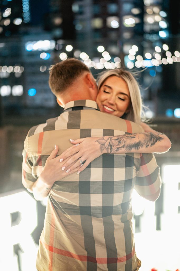 Newly engaged couple embracing at Sparkling Midtown Rooftop with lit windows behind them