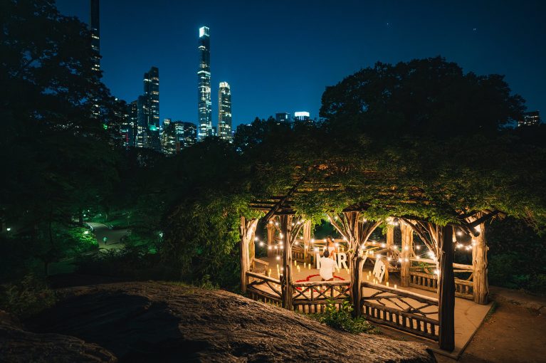 Gazebo in Central Park lit with string lights and candles at night, the Midtown skyline above