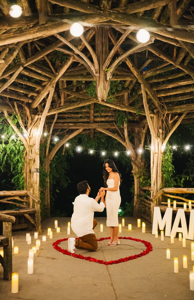 Surprise proposal at Summerhouse at the Dene, Central Park: man on one knee inside a rose petal heart lit by candles