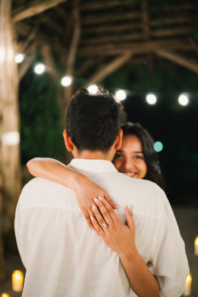Newly engaged couple embracing under string lights at Summerhouse at the Dene, Central Park, her engagement ring visible