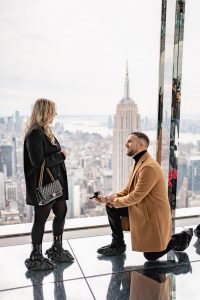 Proposal at SUMMIT One Vanderbilt: man in a camel coat on one knee against the mirrored glass, the Empire State Building behind