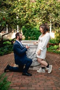 Proposal in the cloister garden of The Met Cloisters: man in a navy suit on one knee by the stone wellhead, brick paving and columns around them