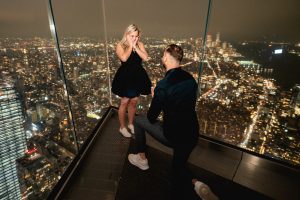 Night proposal at The Edge in Hudson Yards: man on one knee at the glass rail, city lights spread out far below, woman with her hands to her face