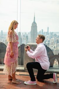Proposal at Top of the Rock: woman in a pink ruffled dress, man on one knee, the Empire State Building framed between them