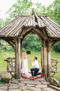 Man proposing on one knee inside a rose petal heart at Wagner Cove in Central Park