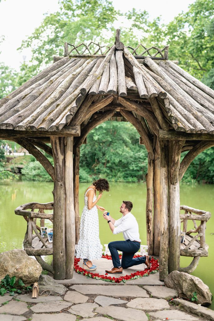 Man proposing on one knee inside a rose petal heart at Wagner Cove in Central Park