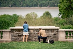 Proposal at Wave Hill: man on one knee on the lawn by the stone wall, the Hudson and the Palisades beyond, a small dog beside them